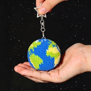 May include: A crocheted Earth keychain with blue oceans and green and yellow continents. The globe is suspended from a silver star-shaped charm. The background is black with white star-like specks.