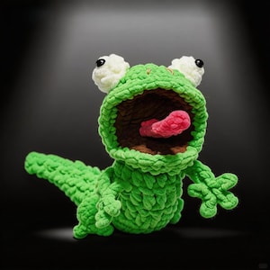 May include: A handmade green crochet frog with a wide open mouth, pink tongue, and white accents. The frog has large, round eyes and is posed with its arms outstretched. The frog is made of a soft, textured yarn.