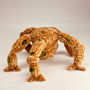 May include: A plush, handmade spider toy in shades of tan and brown, with green accents. The spider has a rounded body and eight articulated legs. The soft, textured material gives it a cozy appearance, suitable for a child's room or as a decorative item.