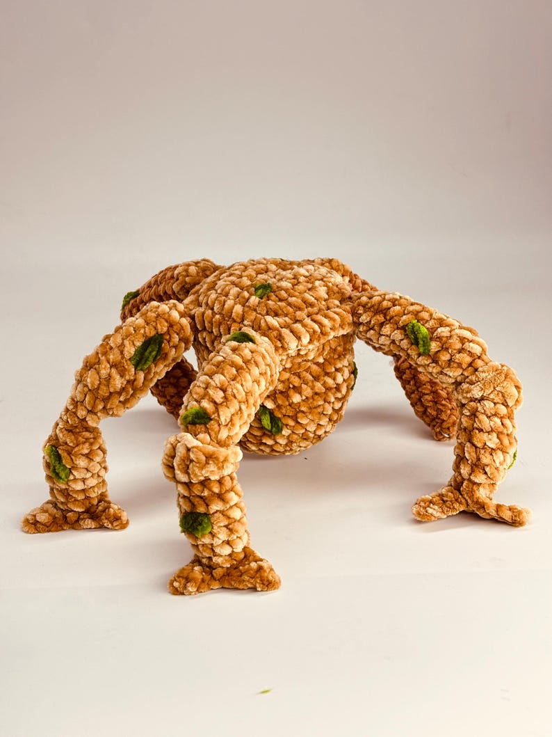 May include: A plush, handmade spider toy in shades of tan and brown, with green accents. The spider has a rounded body and eight articulated legs. The soft, textured material gives it a cozy appearance, suitable for a child's room or as a decorative item.