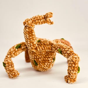 May include: A plush, brown, crocheted creature with four legs and an arm raised in the air. The creature has green accents and a textured surface. The toy is set against a white background.