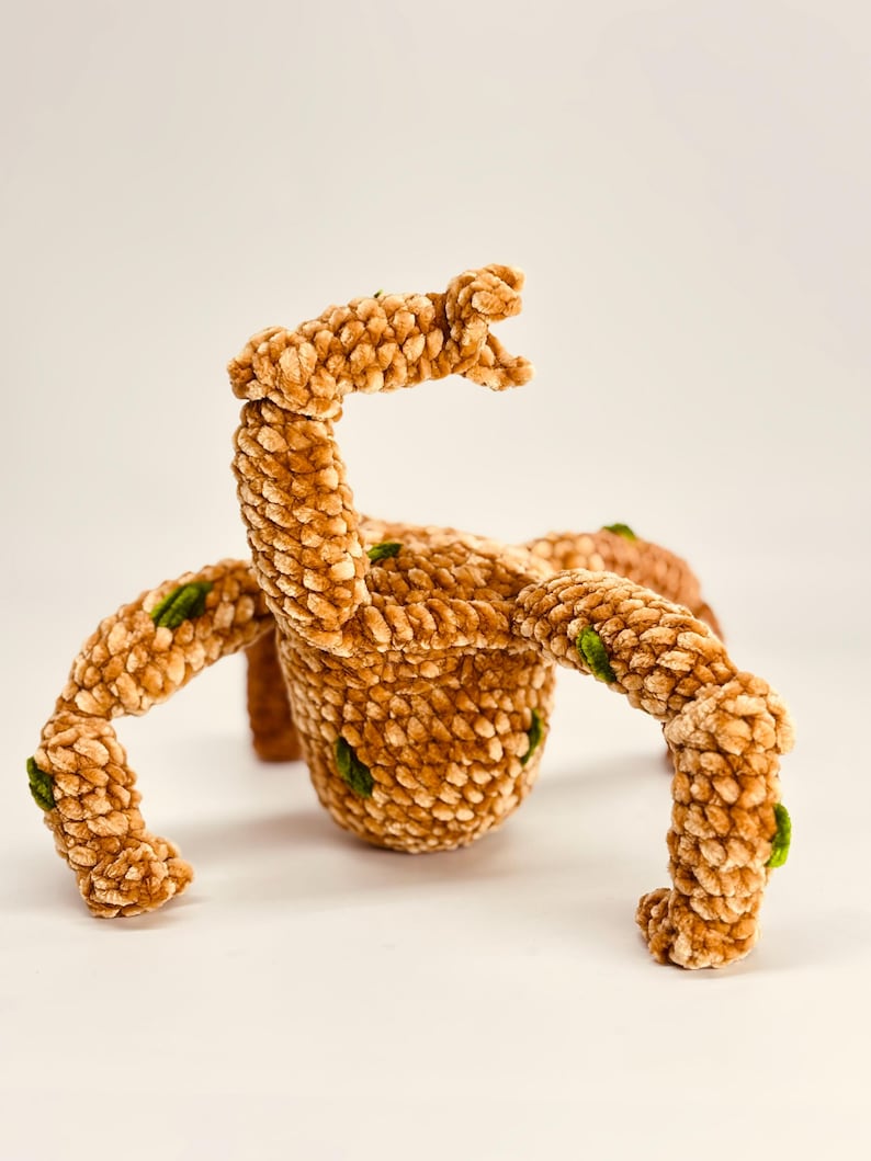May include: A plush, brown, crocheted creature with four legs and an arm raised in the air. The creature has green accents and a textured surface. The toy is set against a white background.