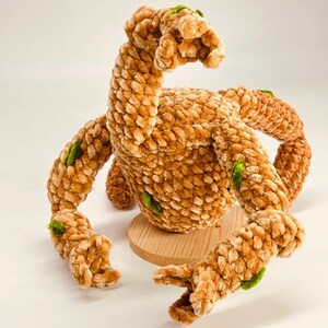 May include: A plush, brown, handmade creature with multiple arms and a curved neck. The soft, textured body has green accents. It sits on a small, round wooden base. The toy is set against a white background.