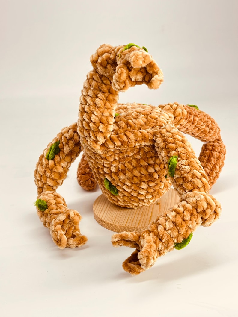 May include: A plush, brown, handmade creature with multiple arms and a curved neck. The soft, textured body has green accents. It sits on a small, round wooden base. The toy is set against a white background.