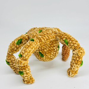 May include: A plush, handmade spider toy in shades of tan and brown, with green accents. The spider has a rounded body and eight legs, each with a textured, velvety appearance. The toy is designed to be soft and cuddly.