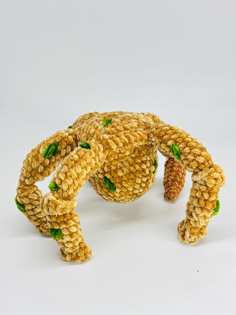 May include: A plush, handmade spider toy in shades of tan and brown, with green accents. The spider has a rounded body and eight legs, each with a textured, velvety appearance. The toy is designed to be soft and cuddly.