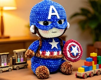 May include: A crocheted plush doll resembling Captain America, featuring a blue textured head with a white letter A, a blue body with a white star, and a red, white, and blue circular shield. The doll has large black eyes and pink cheeks.