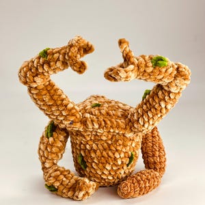 May include: A handmade plush toy in a crab-like design. The toy is made of a soft, textured, light brown material with green accents. The crab has two arms raised and two legs. The toy is sitting on a white surface.