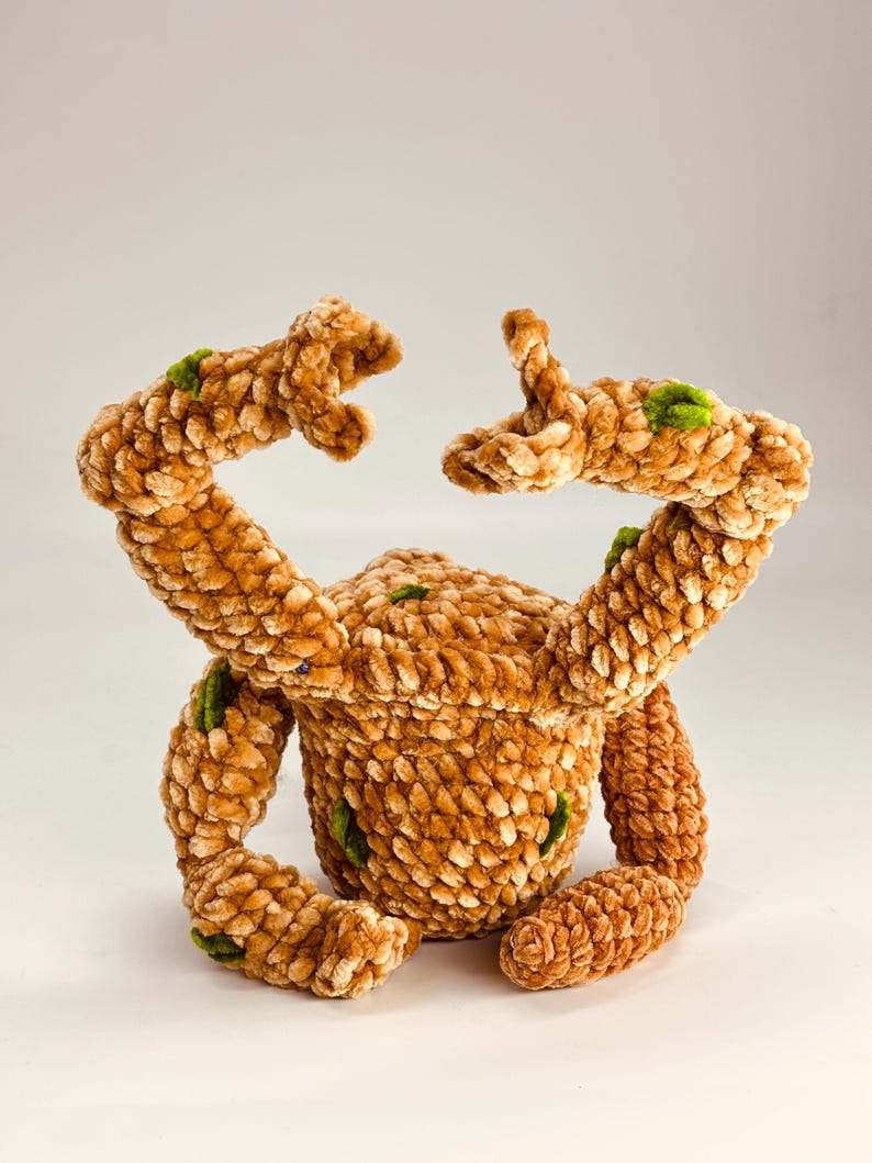 May include: A handmade plush toy in a crab-like design. The toy is made of a soft, textured, light brown material with green accents. The crab has two arms raised and two legs. The toy is sitting on a white surface.