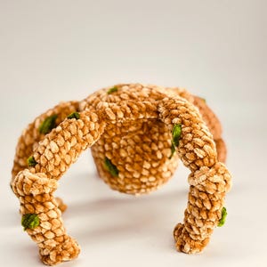May include: A handmade, plush spider toy in shades of tan and brown, with textured, segmented legs and body. Small green accents are visible on the legs. The toy is designed to sit on a surface.