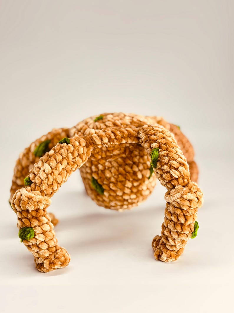 May include: A handmade, plush spider toy in shades of tan and brown, with textured, segmented legs and body. Small green accents are visible on the legs. The toy is designed to sit on a surface.