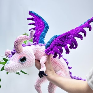 Puede incluir: Un dragón de ganchillo hecho a mano en rosa suave, morado y azul. El peluche presenta alas grandes y detalladas, cuernos y una larga cola. El dragón está sostenido por una mano. Los pies y las garras del dragón son negros.