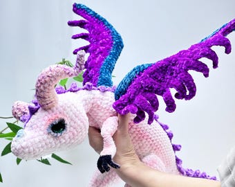 May include: A handmade crochet dragon in soft pink, purple, and blue. The plush toy features large, detailed wings, horns, and a long tail. The dragon is held up by a person's hand. The dragon's feet and claws are black.
