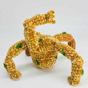 May include: A handmade, plush, tan-colored creature with multiple articulated limbs and a central body. The soft, textured toy features small green accents and is set against a white background. The toy is designed to resemble a fantastical creature.