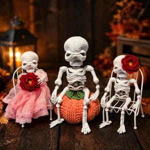 Op de afbeelding: Drie gehaakte witte skeletfiguren. Eén zit op een gebreide oranje pompoen, terwijl twee op witte draadstoelen zitten. Eén skelet draagt een roze kanten jurk met een rode bloem, en een ander heeft een rode bloem aan het hoofd bevestigd.