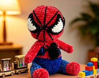 May include: A plush crochet doll featuring a red and blue color scheme with a large round head, white eyes outlined in black, and a black spider emblem on the chest. The doll has a textured, soft appearance and is seated in a forward-facing position.
