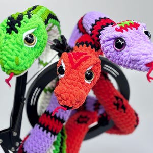 Peut inclure: Trois sculptures de serpents crochet&eacute;s aux couleurs vives. L'un est vert avec des accents noirs, un autre est rouge et le troisi&egrave;me est violet avec des rayures rouges et noires. Chaque serpent a de grands yeux expressifs et une langue rouge.