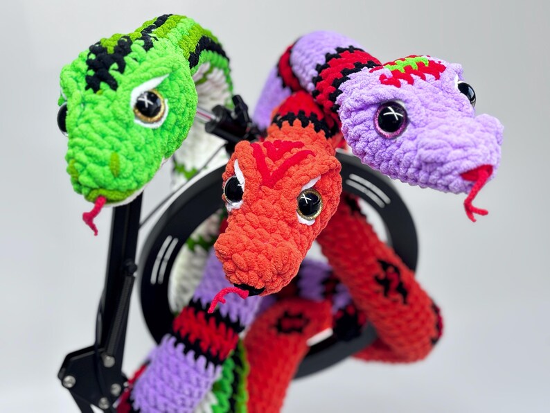 Peut inclure: Trois sculptures de serpents crochet&eacute;s aux couleurs vives. L'un est vert avec des accents noirs, un autre est rouge et le troisi&egrave;me est violet avec des rayures rouges et noires. Chaque serpent a de grands yeux expressifs et une langue rouge.