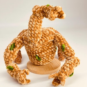 May include: A handmade plush creature in shades of tan and brown, with green accents. The soft, textured toy has a round body, a long neck, and multiple arm-like appendages. It sits on a small, round wooden base.