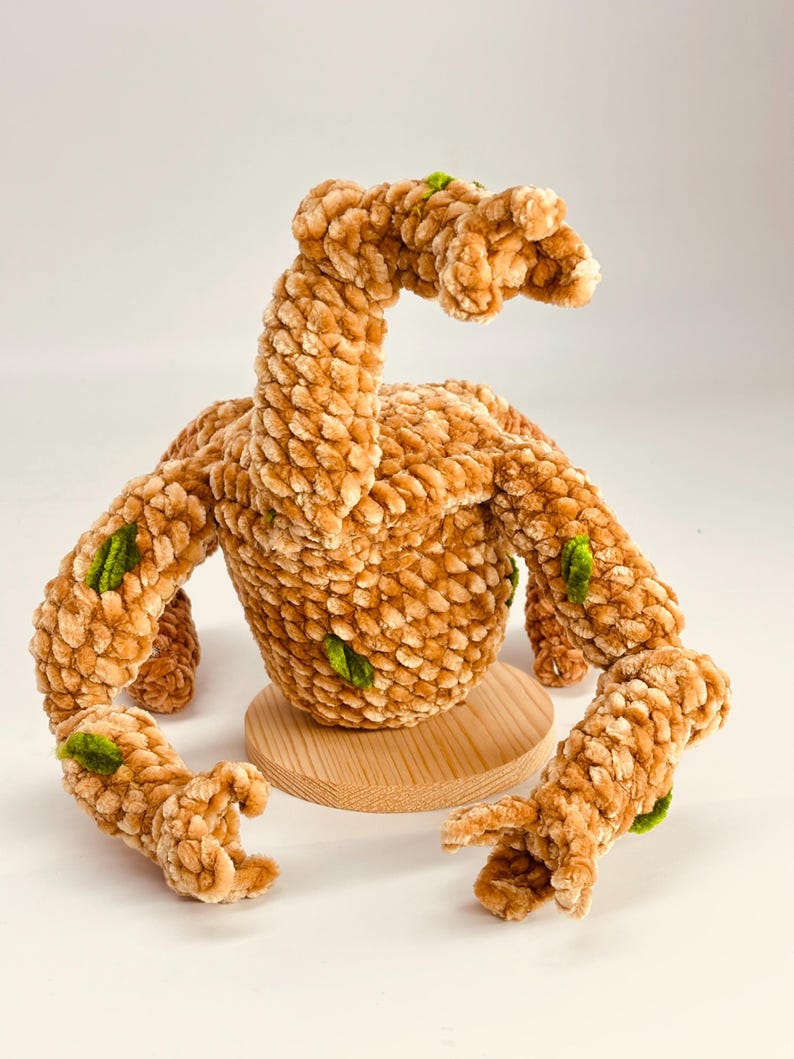 May include: A handmade plush creature in shades of tan and brown, with green accents. The soft, textured toy has a round body, a long neck, and multiple arm-like appendages. It sits on a small, round wooden base.