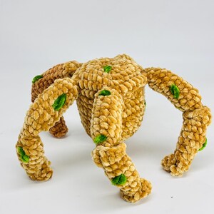 May include: A handmade, plush toy spider in shades of tan and brown, with green accents. The spider has a rounded body and eight segmented legs. The soft, textured material gives the toy a cozy appearance. The toy is designed for children.