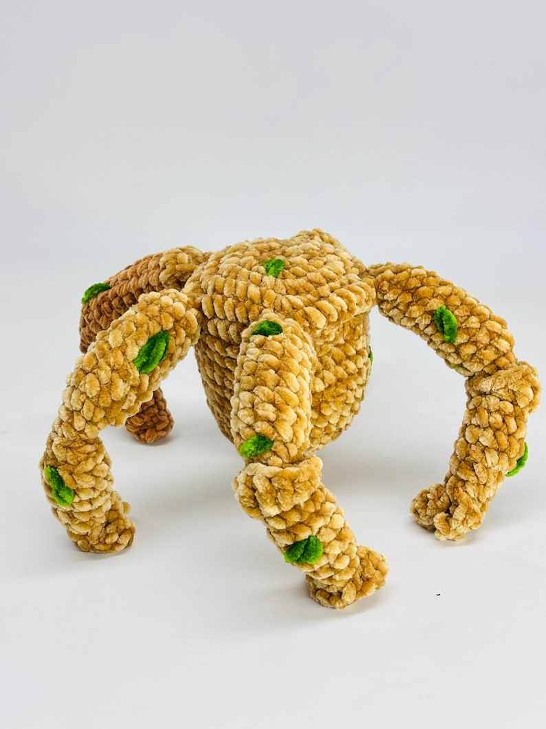 May include: A handmade, plush toy spider in shades of tan and brown, with green accents. The spider has a rounded body and eight segmented legs. The soft, textured material gives the toy a cozy appearance. The toy is designed for children.