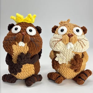 Puede incluir: Dos peluches de castor hechos a mano con ganchillo. Uno es marrón con una corona amarilla, el otro es marrón y beige. Ambos tienen ojos blancos grandes, narices marrones y patas marrones y beige. Están sentados erguidos.