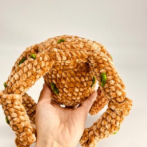 May include: A plush, brown crocheted toy with four extended arms and a central body. The toy has a textured, fuzzy appearance with small green accents. The toy is held in a hand against a white background.