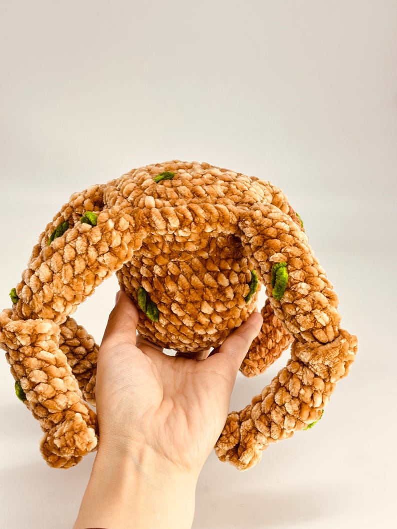 May include: A plush, brown crocheted toy with four extended arms and a central body. The toy has a textured, fuzzy appearance with small green accents. The toy is held in a hand against a white background.