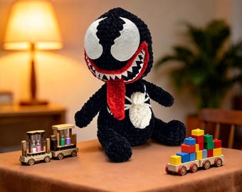 May include: A black crocheted plush toy featuring a large head with white eyes, a wide mouth with sharp white teeth, and a long red tongue. The body has a white spider emblem on the chest. Beside it are a wooden toy train and a block-stacking toy.