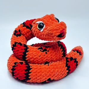 Könnte beinhalten: Ein gehäkeltes Spielzeugschlange in Orange und Schwarz mit schwarzen Augen und einem roten Mund. Die Schlange ist zu einer Spirale zusammengerollt.