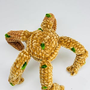 May include: A plush, handmade toy in the shape of a star or creature, with a textured, light brown body and darker brown arms. Green accents are placed on the arms and body. The toy is made of soft, fuzzy material.