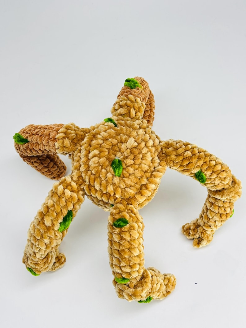 May include: A plush, handmade toy in the shape of a star or creature, with a textured, light brown body and darker brown arms. Green accents are placed on the arms and body. The toy is made of soft, fuzzy material.