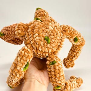 May include: A handmade plush toy in the shape of a creature, featuring a textured, light brown body with green accents. The toy has outstretched arms and legs, and is made of a soft, fuzzy material.
