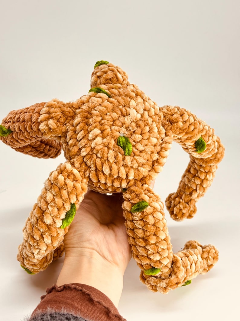 May include: A handmade plush toy in the shape of a creature, featuring a textured, light brown body with green accents. The toy has outstretched arms and legs, and is made of a soft, fuzzy material.