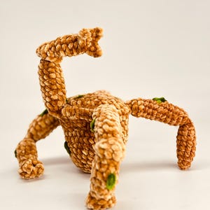 May include: A plush, brown, crocheted creature with multiple legs and an arm raised in the air. The soft toy has green accents and a textured surface, set against a plain white background.