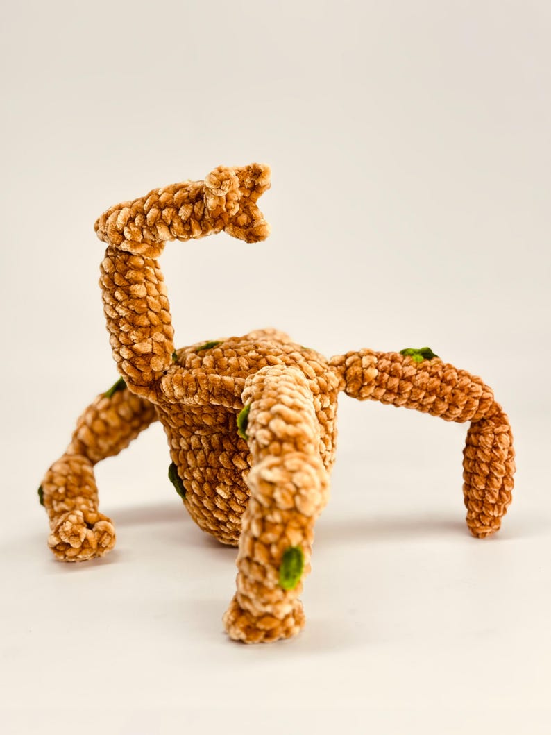 May include: A plush, brown, crocheted creature with multiple legs and an arm raised in the air. The soft toy has green accents and a textured surface, set against a plain white background.