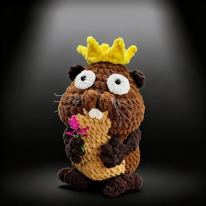 Puede incluir: Una muñeca de castor hecha a mano, con una corona amarilla y una flor rosa. El castor es principalmente marrón, con detalles en marrón claro y oscuro. Tiene grandes ojos blancos con pupilas negras y largos bigotes. Un artículo artesanal caprichoso.
