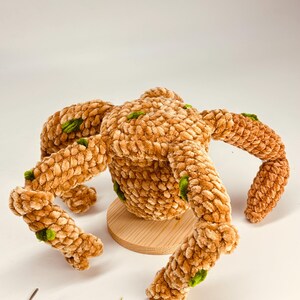 May include: A plush, brown crocheted creature with four legs and a round body, accented with green details. The handmade item is displayed on a small, round wooden base, showcasing its unique texture and design. A crochet hook lies nearby.
