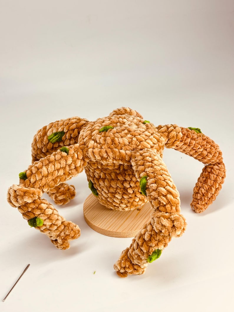 May include: A plush, brown crocheted creature with four legs and a round body, accented with green details. The handmade item is displayed on a small, round wooden base, showcasing its unique texture and design. A crochet hook lies nearby.