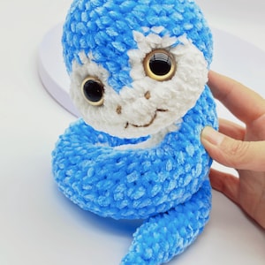 Puede incluir: Un juguete de serpiente de ganchillo azul y blanco con ojos negros grandes y brillantes y una boca sonriente. La serpiente de peluche está hecha de hilo suave y texturizado y está enrollada, con la cabeza erguida. El juguete está sostenido por una mano.
