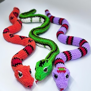 Op de afbeelding: Drie gehaakte slangen speelgoed in levendige kleuren. Eén is rood met zwarte markeringen, een andere is groen met zwarte strepen, en de derde is paars met rode en zwarte strepen. Elke slang heeft een gedetailleerde kop met ogen en een rode tong.