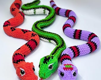 May include: Three colourful crocheted snake toys. One is red with black markings, another is green with black stripes, and the third is purple with red and black stripes. Each snake has a detailed head with eyes and a red tongue.