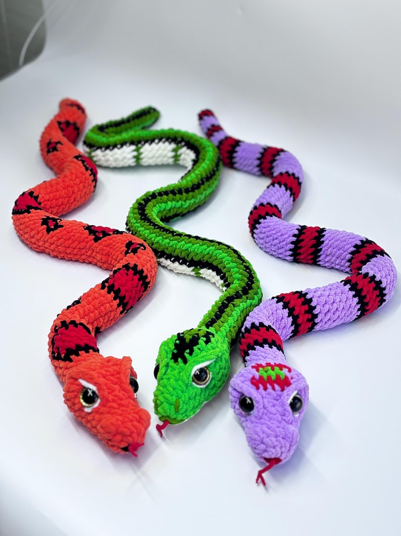 Peut inclure: Trois jouets en forme de serpent crochet&eacute;s aux couleurs vives. L'un est rouge avec des marques noires, un autre est vert avec des rayures noires, et le troisi&egrave;me est violet avec des rayures rouges et noires. Chaque serpent a une t&ecirc;te d&eacute;taill&eacute;e avec des yeux et une langue rouge.
