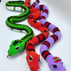 Peut inclure: Trois jouets en forme de serpent crochet&eacute;s aux couleurs vives. L'un est vert avec des accents noirs et blancs, un autre est rouge avec des marques noires et le troisi&egrave;me est violet avec des rayures rouges et noires. Chaque serpent a de grands yeux globuleux et une langue rouge.