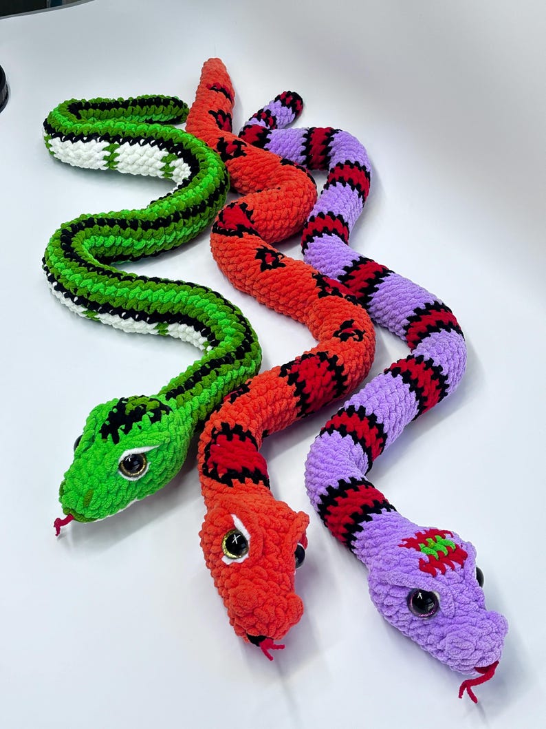 Peut inclure: Trois jouets en forme de serpent crochet&eacute;s aux couleurs vives. L'un est vert avec des accents noirs et blancs, un autre est rouge avec des marques noires et le troisi&egrave;me est violet avec des rayures rouges et noires. Chaque serpent a de grands yeux globuleux et une langue rouge.