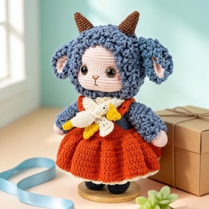 May include: A handmade crochet lamb doll with a blue hood and a red dress. The doll has brown horns, large black eyes, and a white and yellow scarf. It stands on a wooden base and is surrounded by decorative elements.