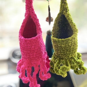 Könnte beinhalten: Zwei handgefertigte Häkel-Pflanzenhalter in leuchtenden Farben. Einer ist leuchtend rosa, der andere olivgrün. Jeder hat einen beutelartigen Körper und mehrere herabhängende Ranken, die einem Oktopus ähneln. Die Halter sind aufgehängt, was darauf hindeutet, dass sie zum Aufhängen bestimmt sind.