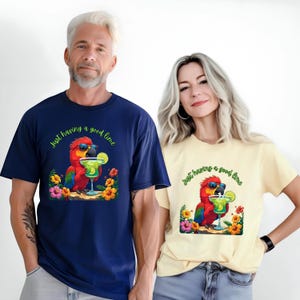 May include: Navy blue and pale yellow t-shirts with a parrot graphic. The parrot wears sunglasses and holds a margarita. The text "Just having a good time" is above the parrot. Floral details surround the image.