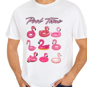 May include: White t-shirt with a graphic of nine pink flamingo pool floats. The words "Pool Time" are written above the floats in a cursive font. The shirt is a casual, summer-themed design.